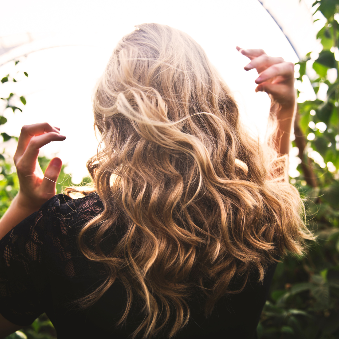 Person with long, wavy blonde hair standing outdoors with greenery in the background