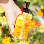 Hand holding a bottle of 'bare flowers' botanical body oil with flowers in the background