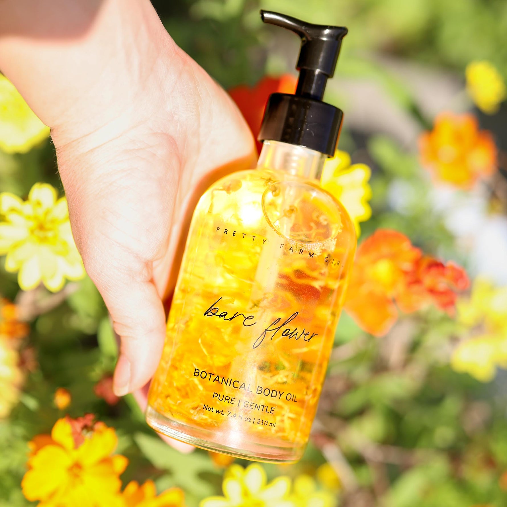 Hand holding a bottle of 'bare flowers' botanical body oil with flowers in the background