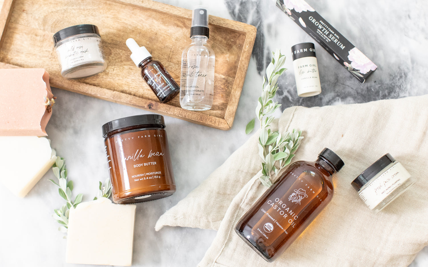 Collection of Pretty Farm Girl's best selling organic skincare products on a marble surface with a wooden tray. Products shown are Wild Rose Probiotic mask, Coffee Bean Eye Lift Serum, Rose Facial Toner, Vanilla bean Body butter, organic castor oil, farm girl vanilla bean lip butter, growth serum, and Fresh Face Cream.