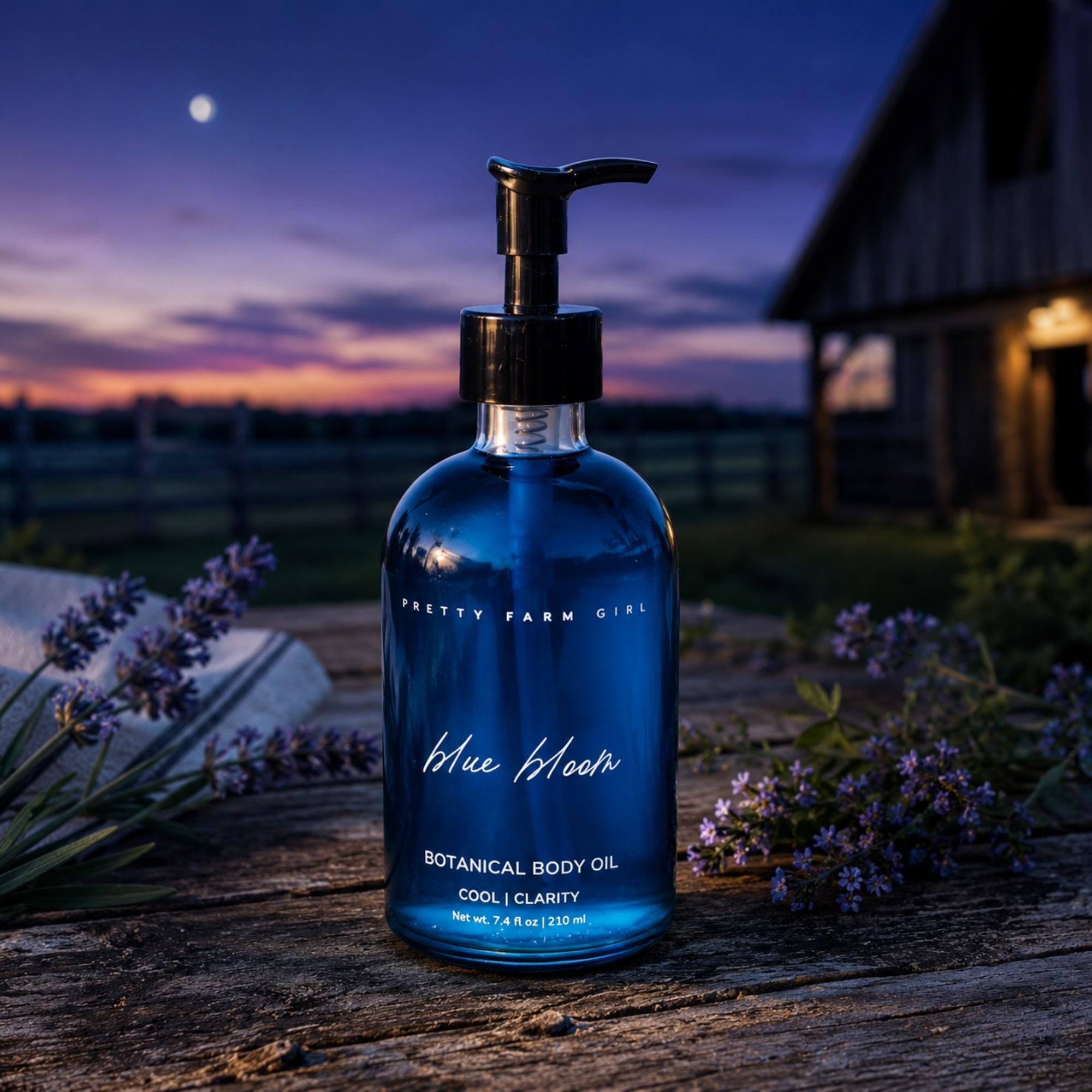 A bottle of Pretty Farm Girl Blue Bloom Botanical Body Oil stands on a rustic wooden surface outdoors at dusk, surrounded by lavender flowers, with a barn and twilight sky in the background.