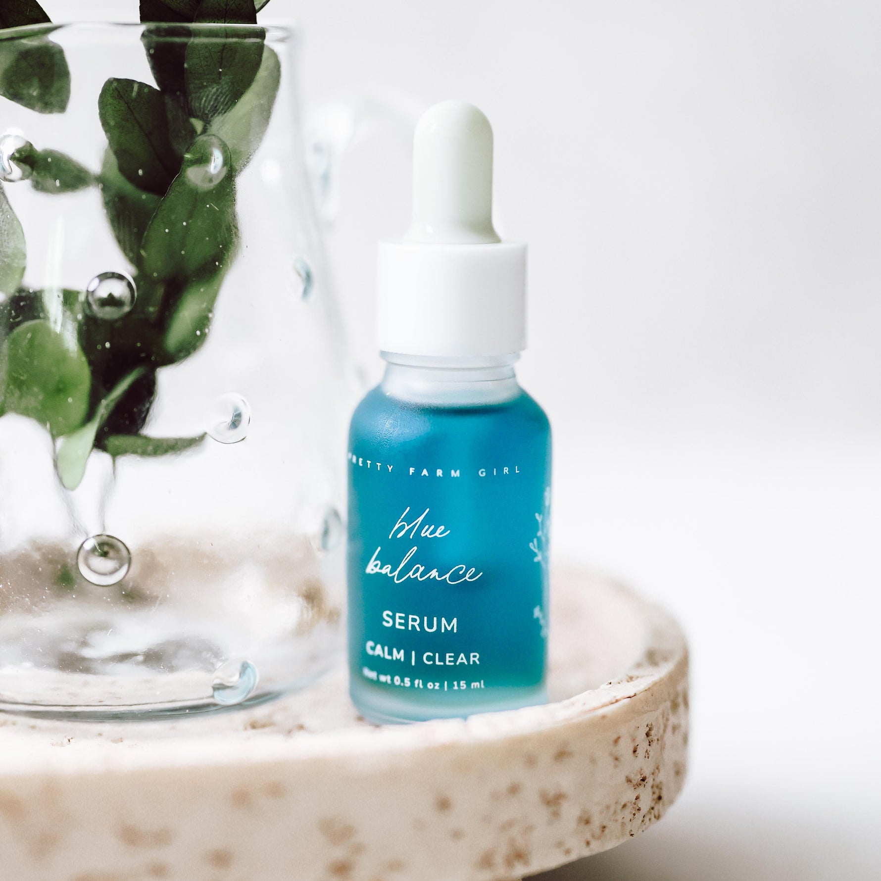 Blue serum bottle with dropper on a white surface next to green leaves
