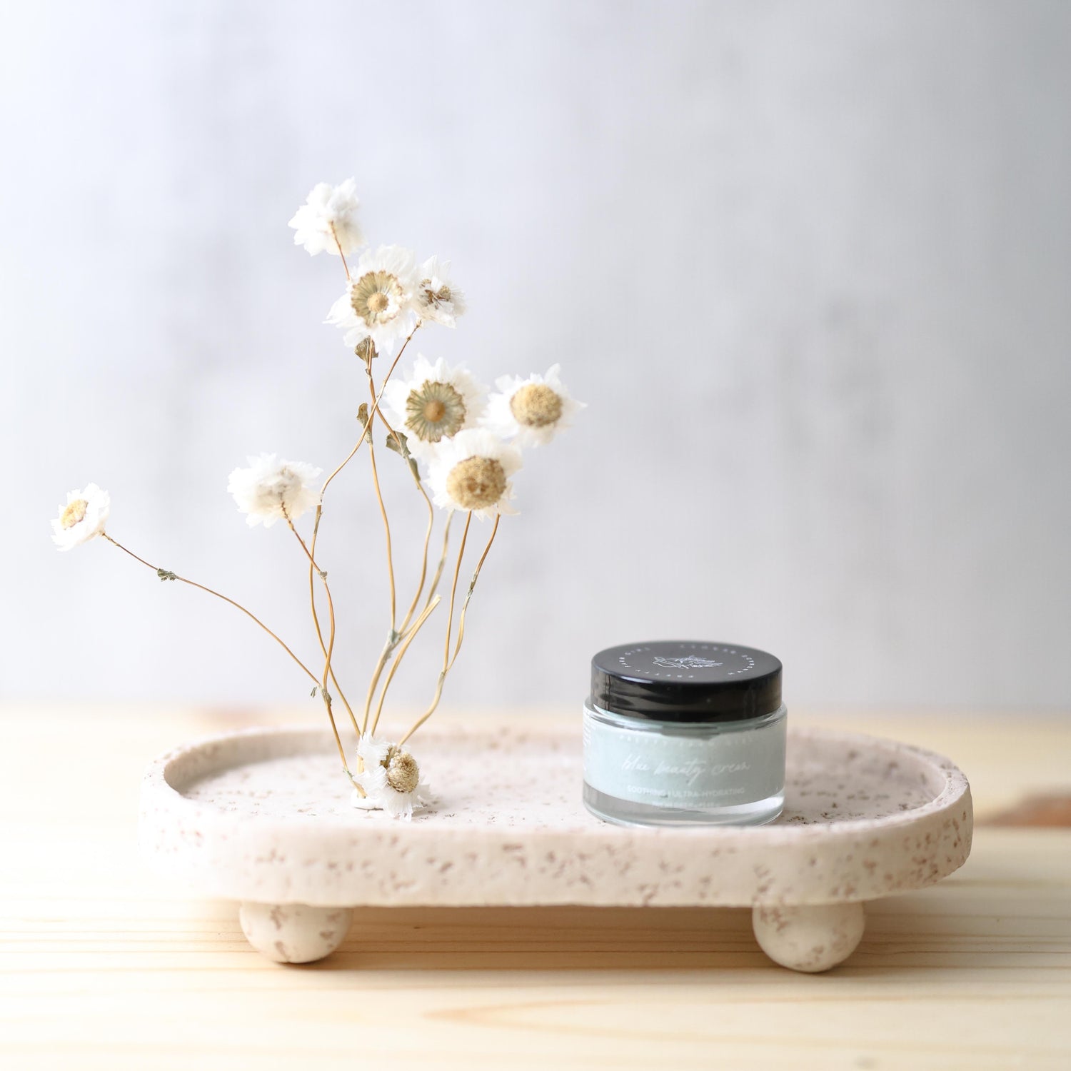 Jar of blue beauty cream on a decorative tray with dried flowers against a light background