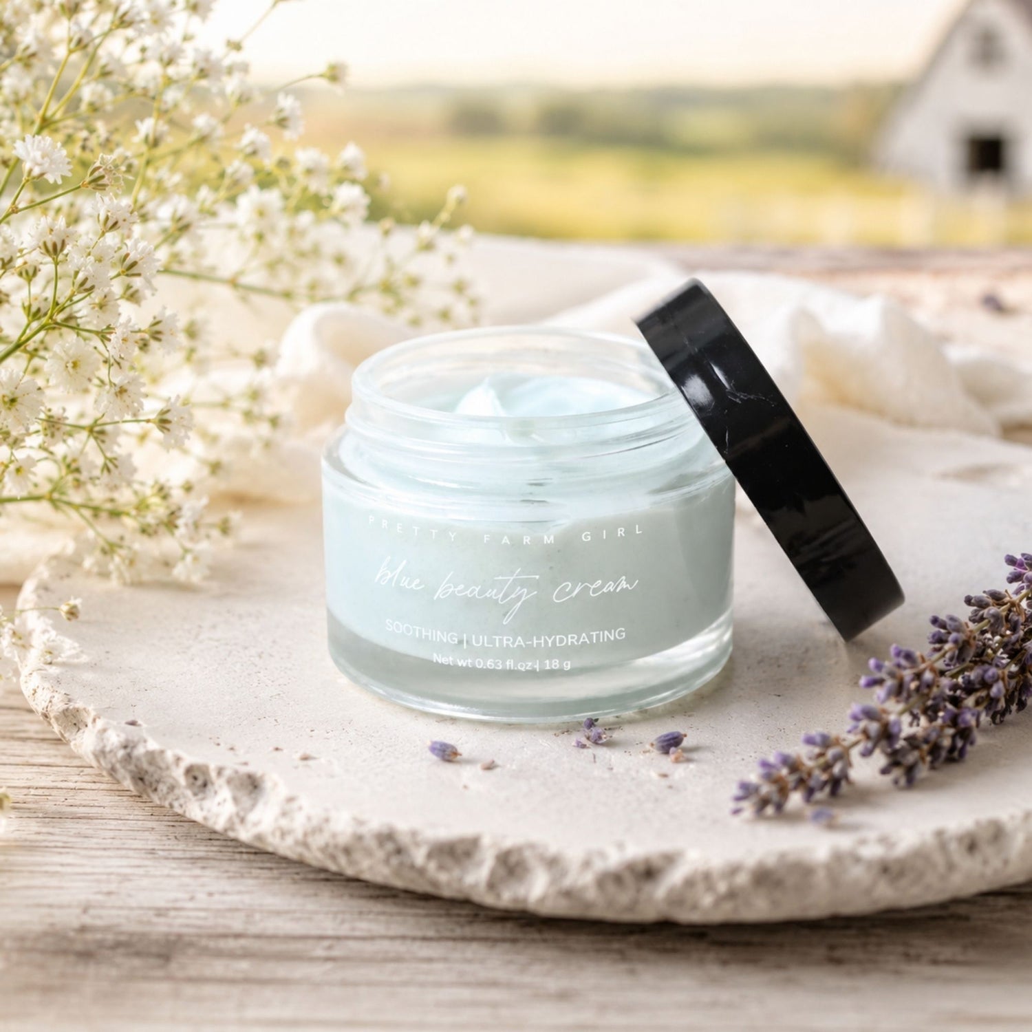 A cosmetic jar labeled 'Blue Beauty Cream' by Pretty Lawn Girl, filled with pale blue cream and set on a stone slab with a black lid leaning beside it. White flowers and lavender sprigs decorate the scene against a softly blurred outdoor background.