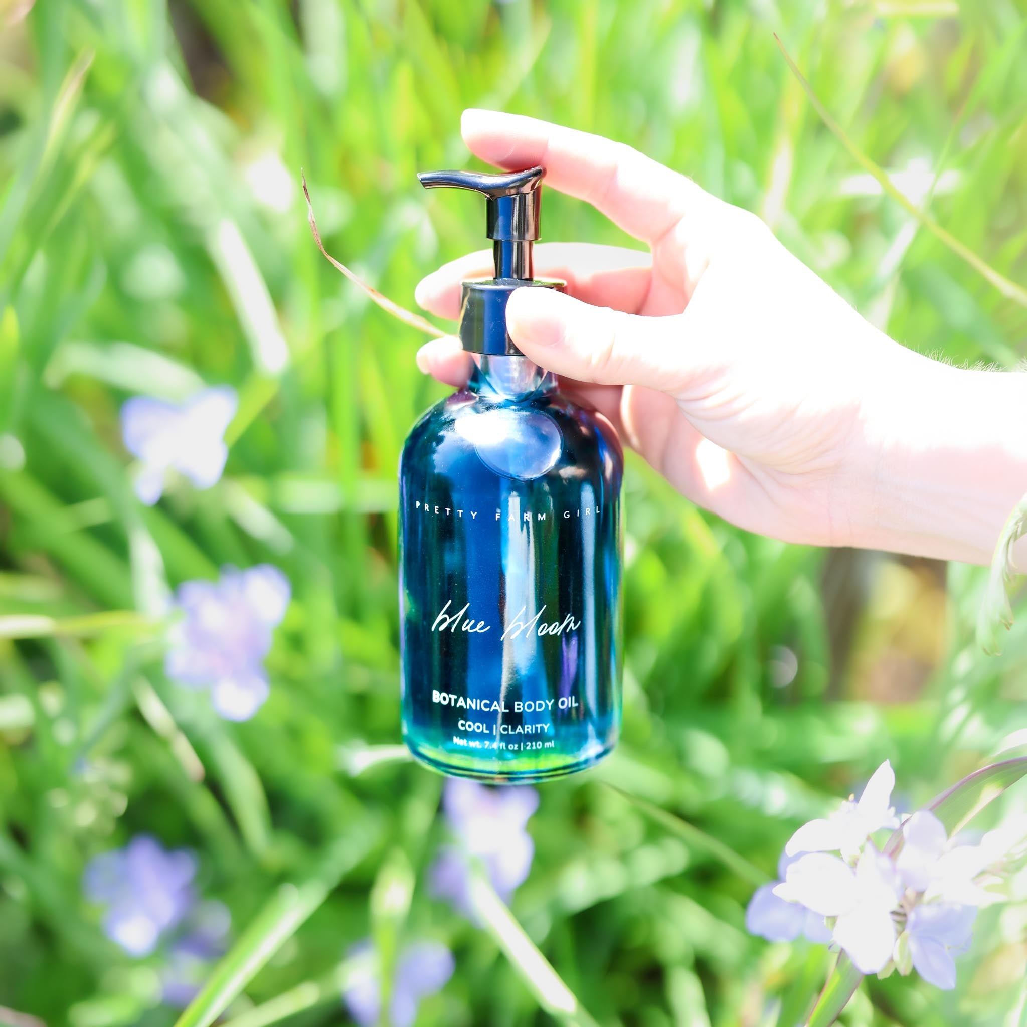 Blue Bloom Botanical Body Oil glass bottle held outdoors among flowers