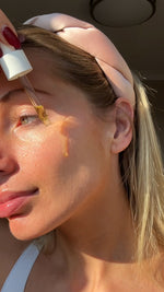 Woman applying dropper of Brightening Vitamin C serum to her cheekbone with a blurred background