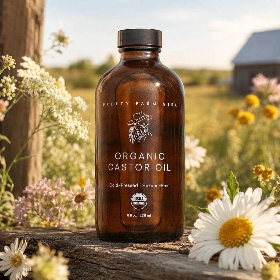 A bottle of Pretty Farm Girl Organic Castor Oil stands on a rustic wooden surface outdoors, surrounded by white and yellow wildflowers, with a blurred farm background.