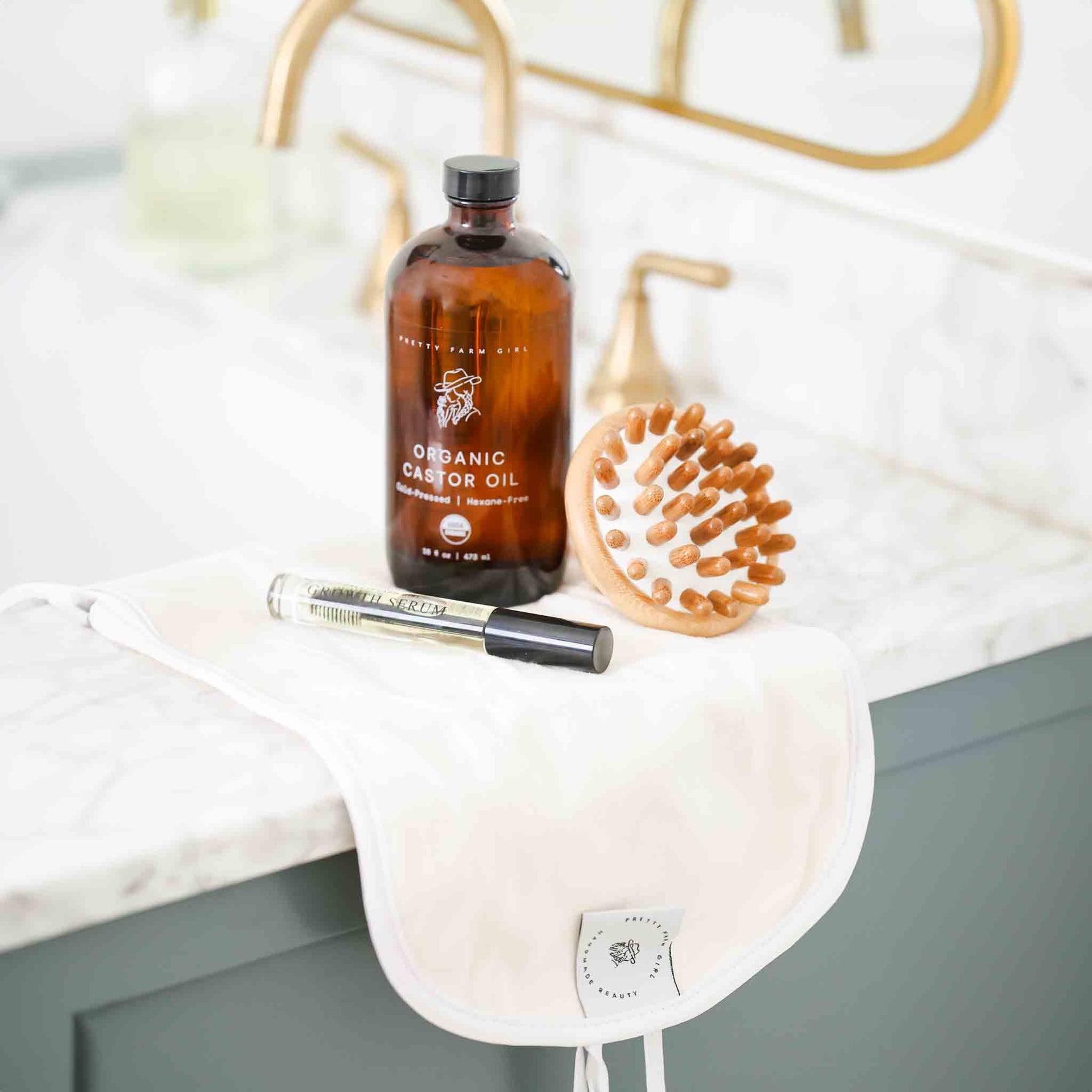 Bottle of organic castor oil bottle, castor oil cloth pack, bamboo scalp massage, and eyelash serum on a bathroom sink counter