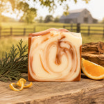 A rectangular bar of Citrus Cedarwood soap with a cream and tan marbled pattern stands on a rustic wooden surface outdoors. The soap is styled with an orange peel curl, a slice of orange, and greenery, with a blurred farm background showing a barn and fence.