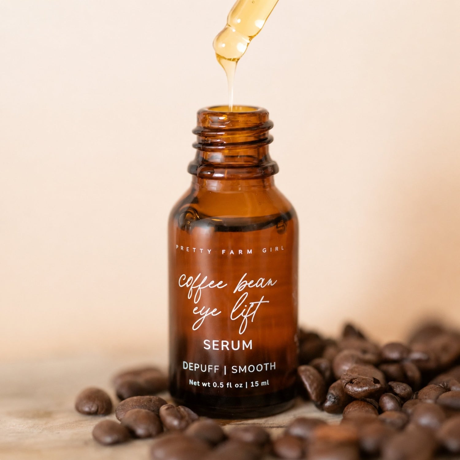 An amber glass bottle labeled 'Pretty Farm Girl Coffee Bean Eye Lift Serum' with a golden liquid being dispensed above it, surrounded by coffee beans on a light beige background.
