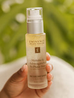 Hand holding a bottle of Eminence skincare product against a blurred green background