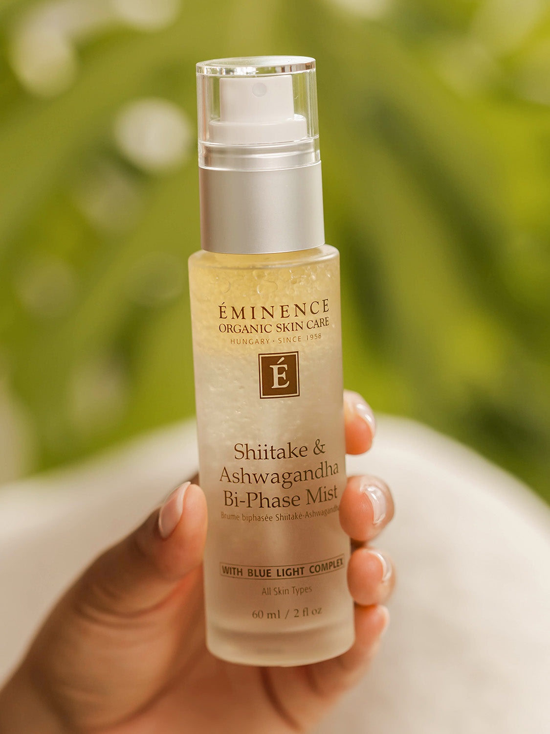 Hand holding a bottle of Eminence skincare product against a blurred green background