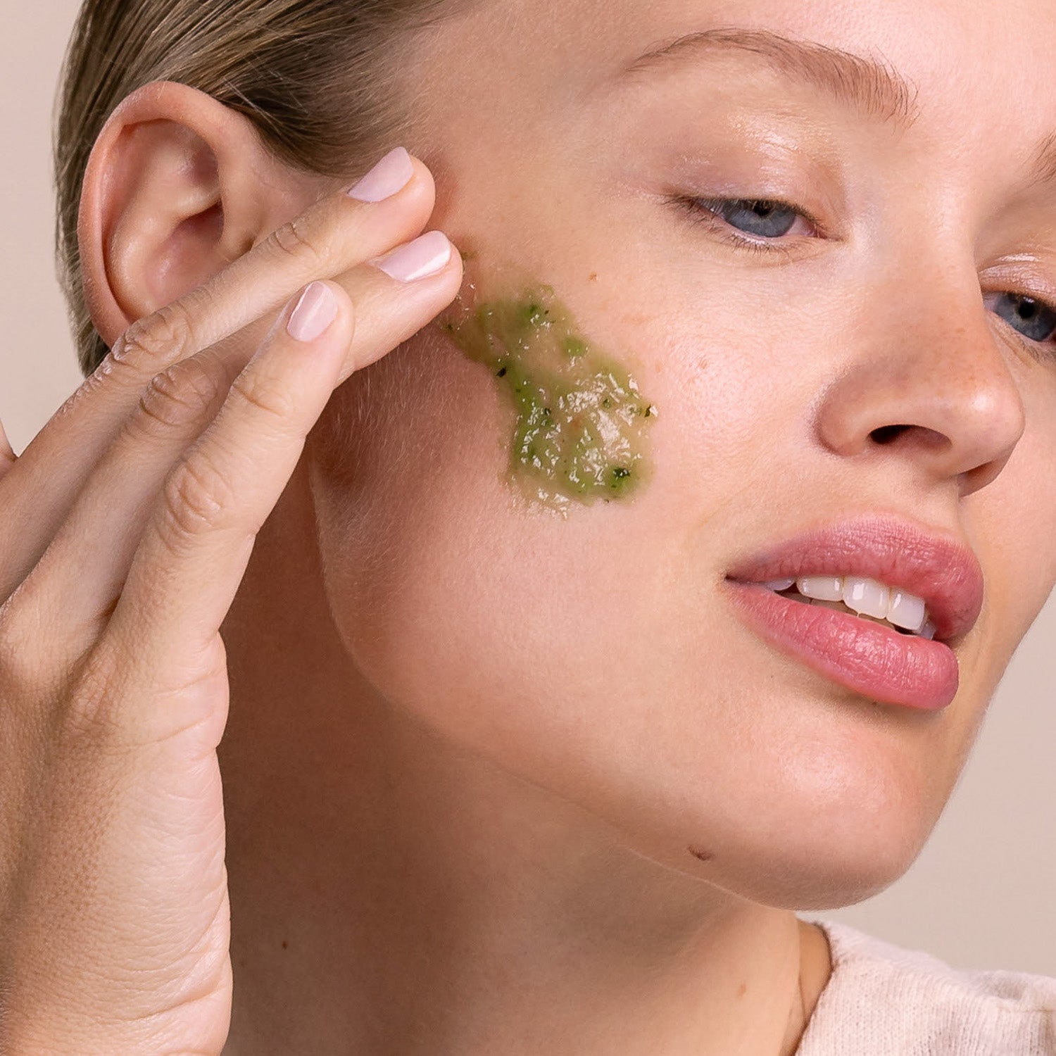 Woman applying green facial mask to her cheek with a neutral background