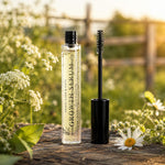 A Pretty Farm Girl tube labeled 'Growth Serum' stands on a wooden surface outdoors, with the black wand removed and placed beside it. White wildflowers and greenery are blurred in the background.