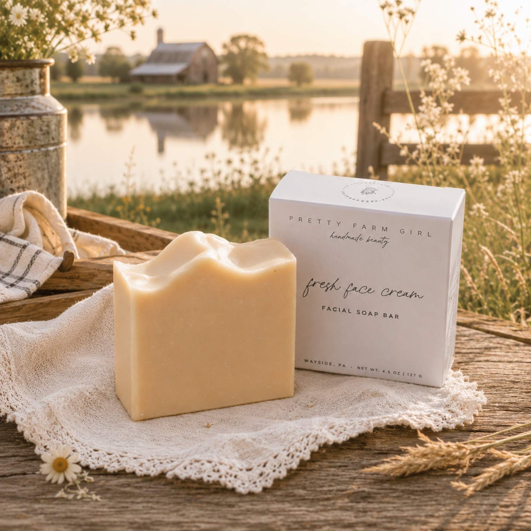 A handmade soap bar and its white packaging box are displayed on a rustic wooden table draped with a lace cloth, set against a warm, golden sunset background of a lake and farm fields. The soap is a warm cream color, and the box is white with black text reading 'PRETTY FARM GIRL handmade beauty' and 'fresh face cream FACIAL SOAP BAR'.