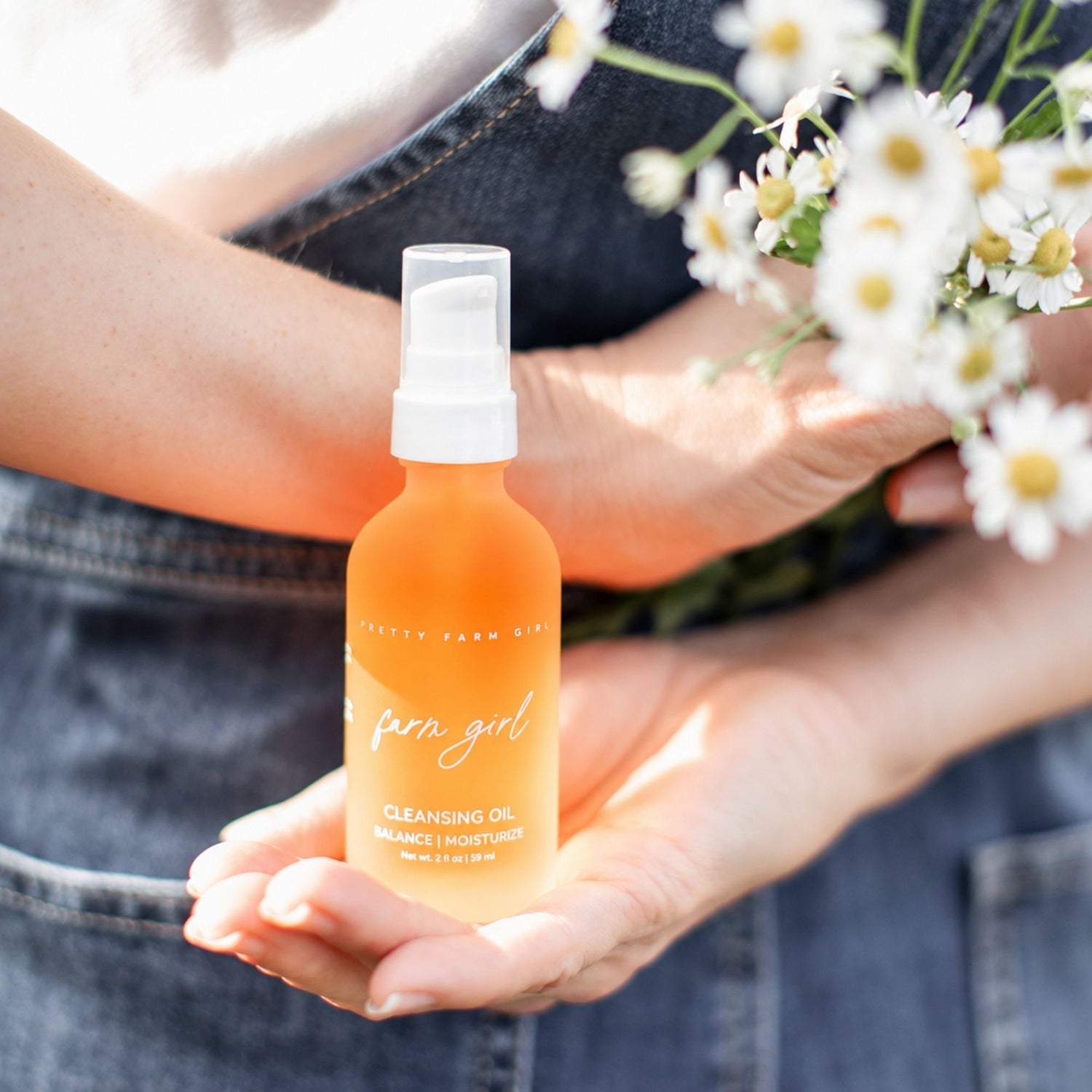 A person in denim overalls holds a bottle of Farm Girl Cleansing Oil in their hands, with white daisies in the background.