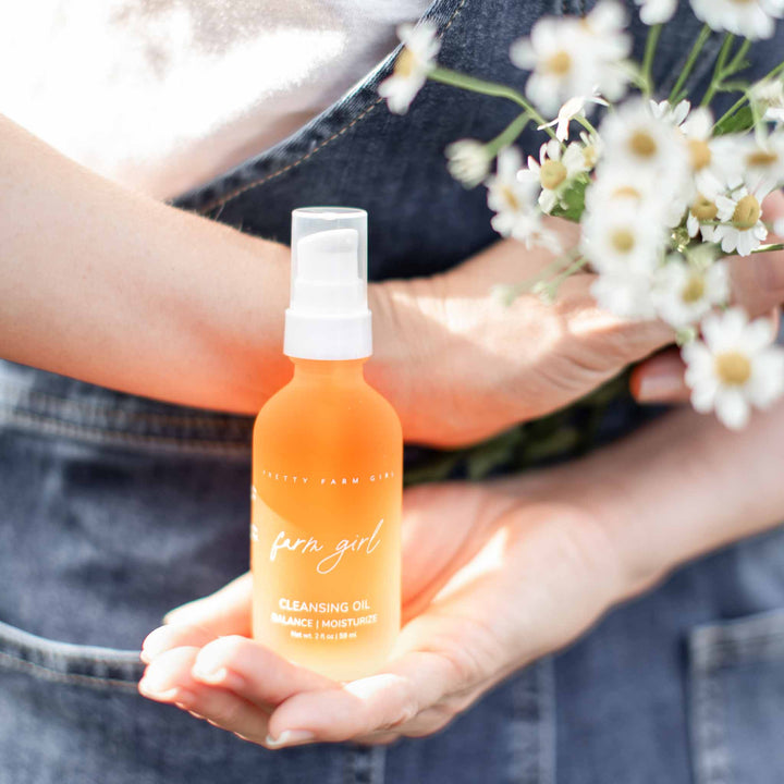 Person holding orange pretty farm girl cleansing oil with a white cap, surrounded by small white flowers.