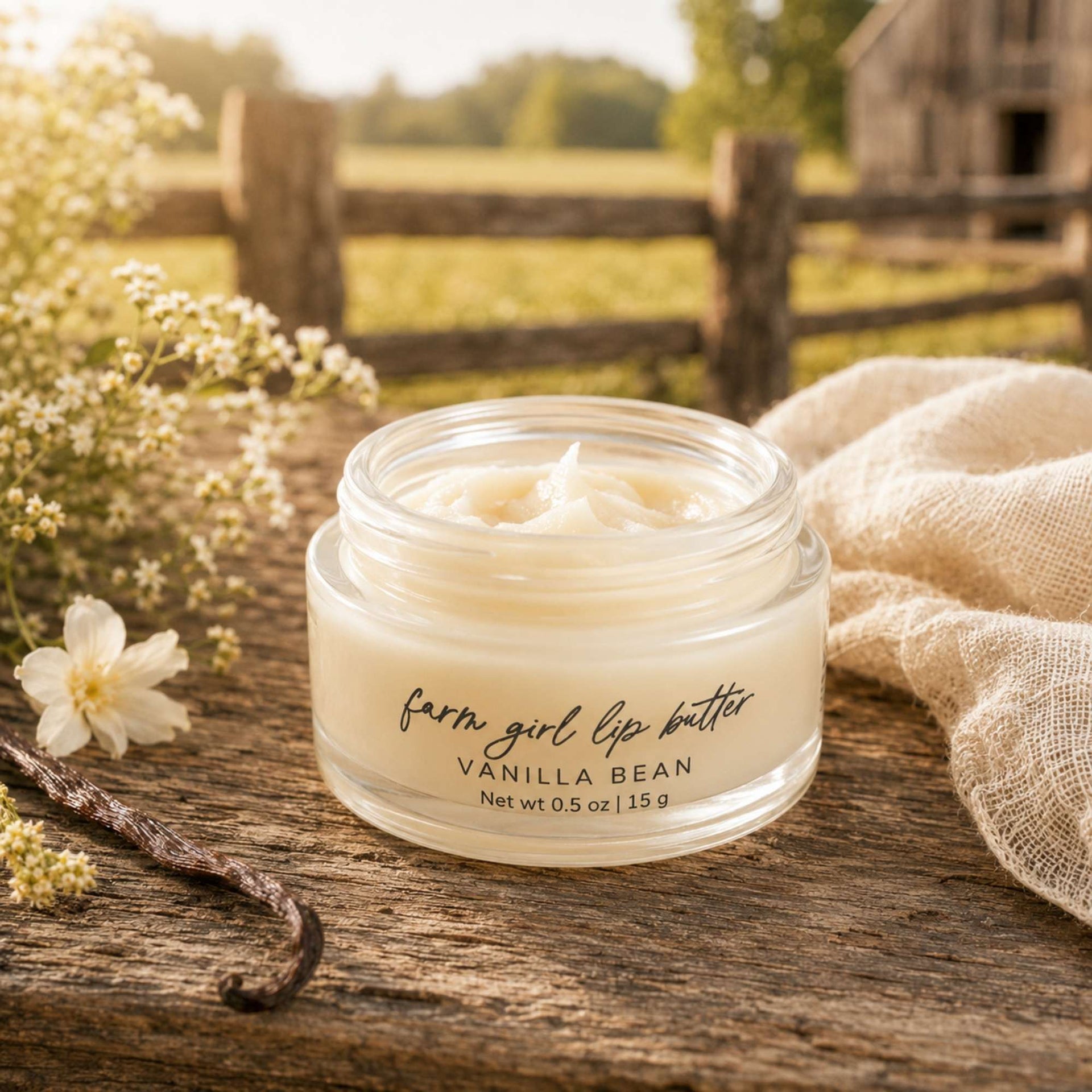 A small clear jar of lip balm labeled 'farm girl lip butter' and 'VANILLA BEAN' sits on a rustic wooden surface outdoors, with flowers, a vanilla bean, and a beige cloth around it and a blurred fence and barn in the background.