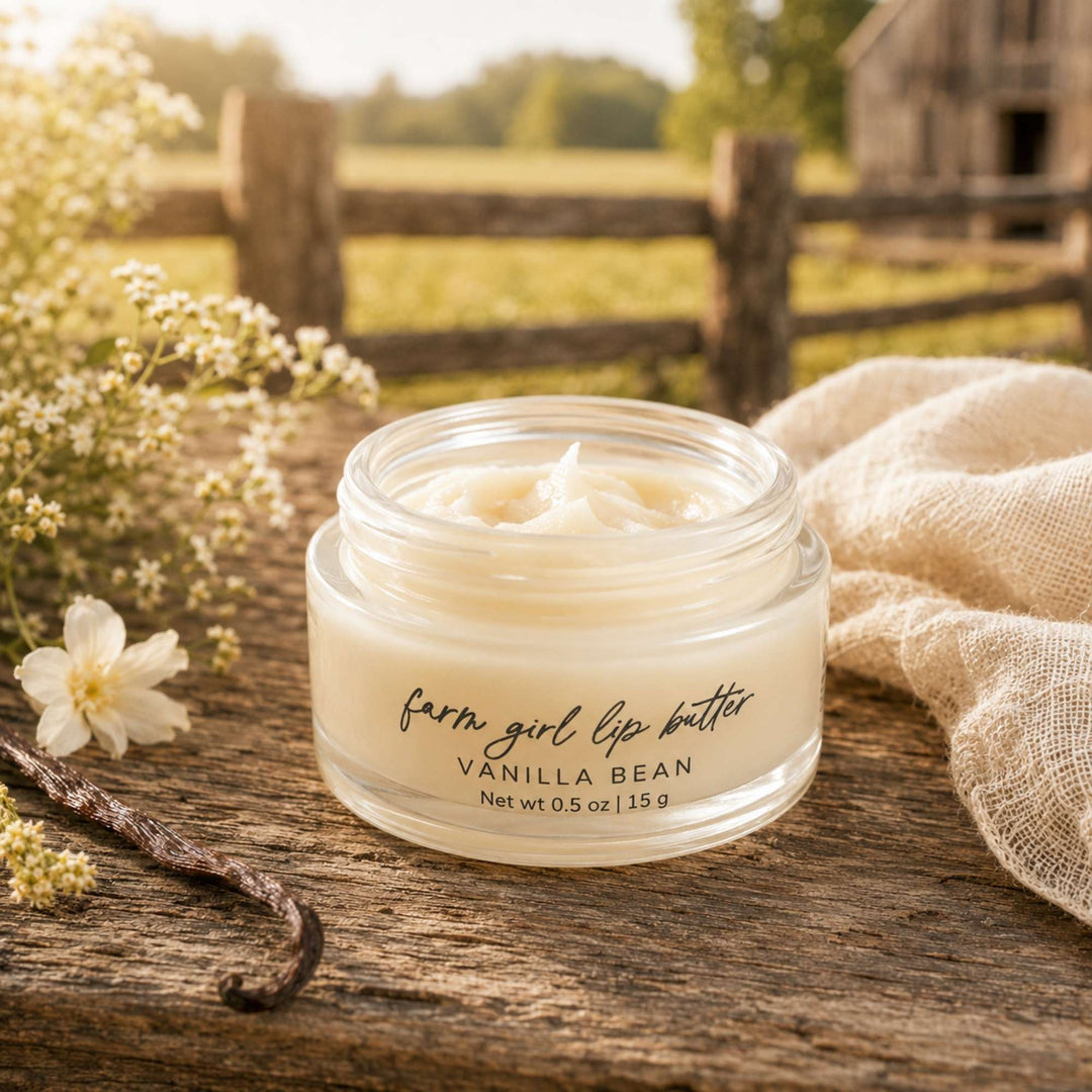 A small clear jar of lip balm labeled 'farm girl lip butter' and 'VANILLA BEAN' sits on a rustic wooden surface outdoors, with flowers, a vanilla bean, and a beige cloth around it and a blurred fence and barn in the background.