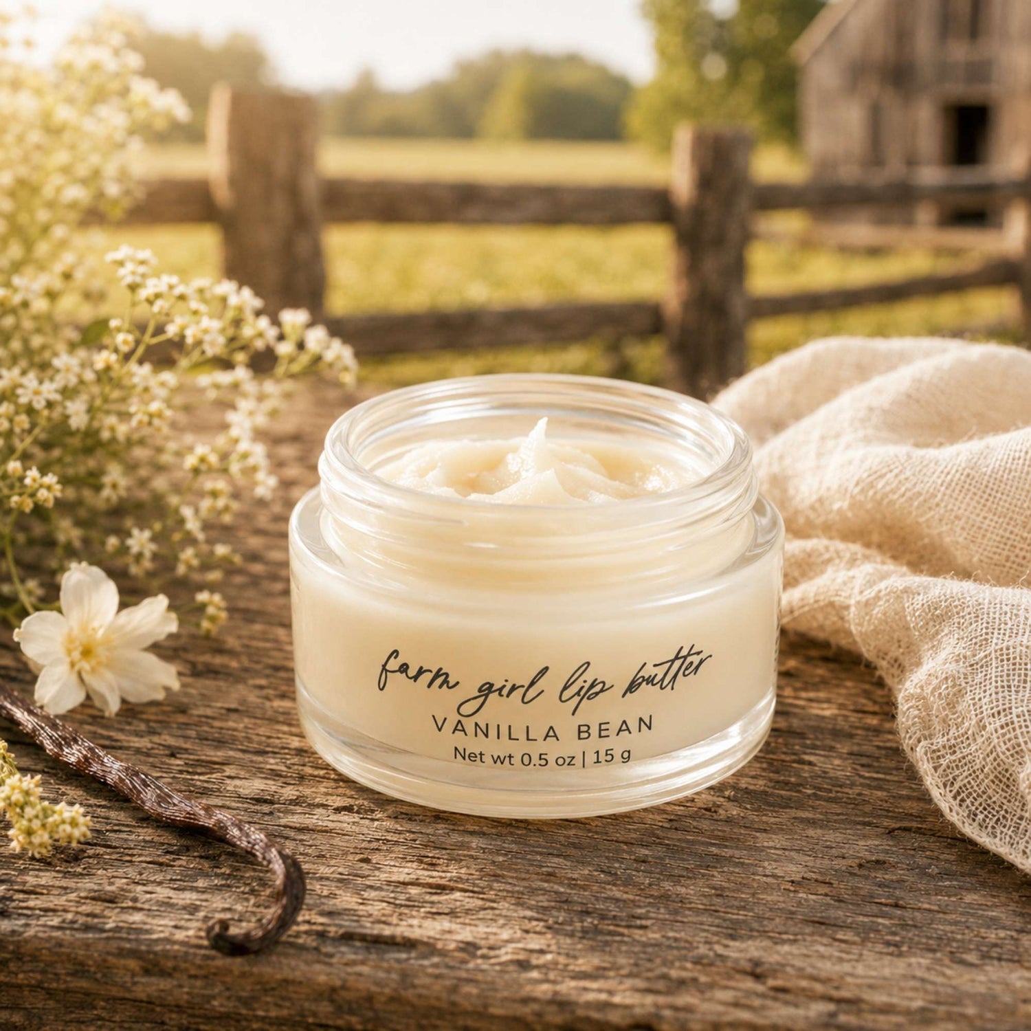 A small clear jar of lip balm labeled 'farm girl lip butter' and 'VANILLA BEAN' sits on a rustic wooden surface outdoors, with flowers, a vanilla bean, and a beige cloth around it and a blurred fence and barn in the background.