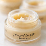 A close-up of a small clear cosmetic jar labeled 'farm girl lip butter VANILLA BEAN' filled with a pale creamy yellow lip balm, with two more jars blurred in the background.