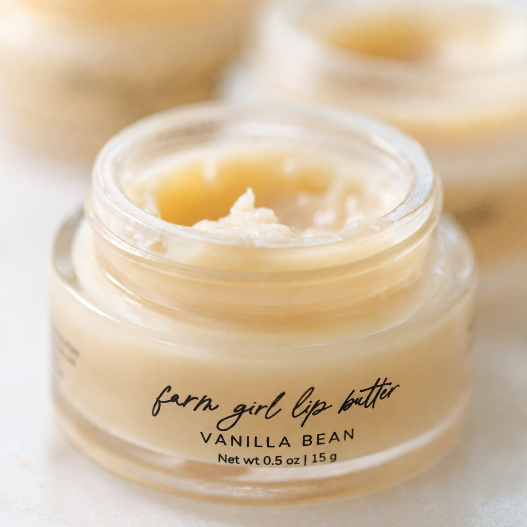 A close-up of a small clear cosmetic jar labeled 'farm girl lip butter VANILLA BEAN' filled with a pale creamy yellow lip balm, with two more jars blurred in the background.