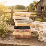 A stack of eight artisanal handmade soap bars in various colors and textures, styled on a rustic wooden table with a linen cloth and baby's breath flowers, set against a blurred background of a barn and field.