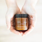 A person's hands holding a small amber glass jar of Pretty Farm Girl 'Tallow + Honey Cream' against a soft, warm beige background.