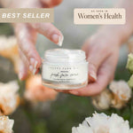 Person holding a 0.78oz glass jar of Pretty Farm Girl's beef tallow Original Fresh Face Cream with flowers in the background.