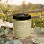 A jar of Pretty Farm Girl cleansing grains in the green tea + coconut milk scent, set on a rustic wooden table with eucalyptus leaves and a small dish of powder, with a blurred outdoor garden background.