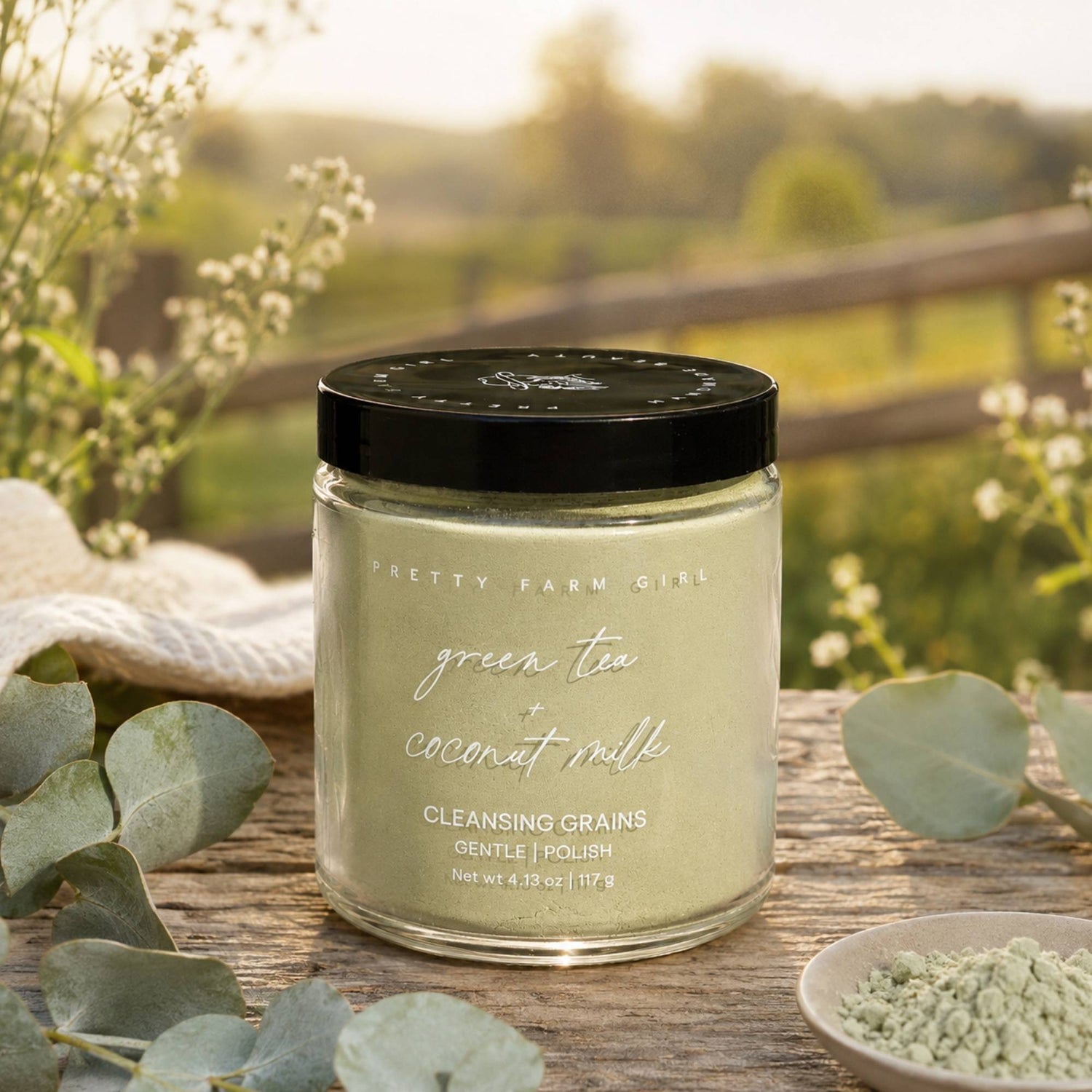 A jar of Pretty Farm Girl cleansing grains in the green tea + coconut milk scent, set on a rustic wooden table with eucalyptus leaves and a small dish of powder, with a blurred outdoor garden background.