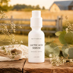 A white bottle of Pretty Farm Girl Lactic Acid Serum stands on a rustic wooden surface outdoors, surrounded by eucalyptus leaves, small white flowers, and a soft beige cloth, with a blurred farm background.