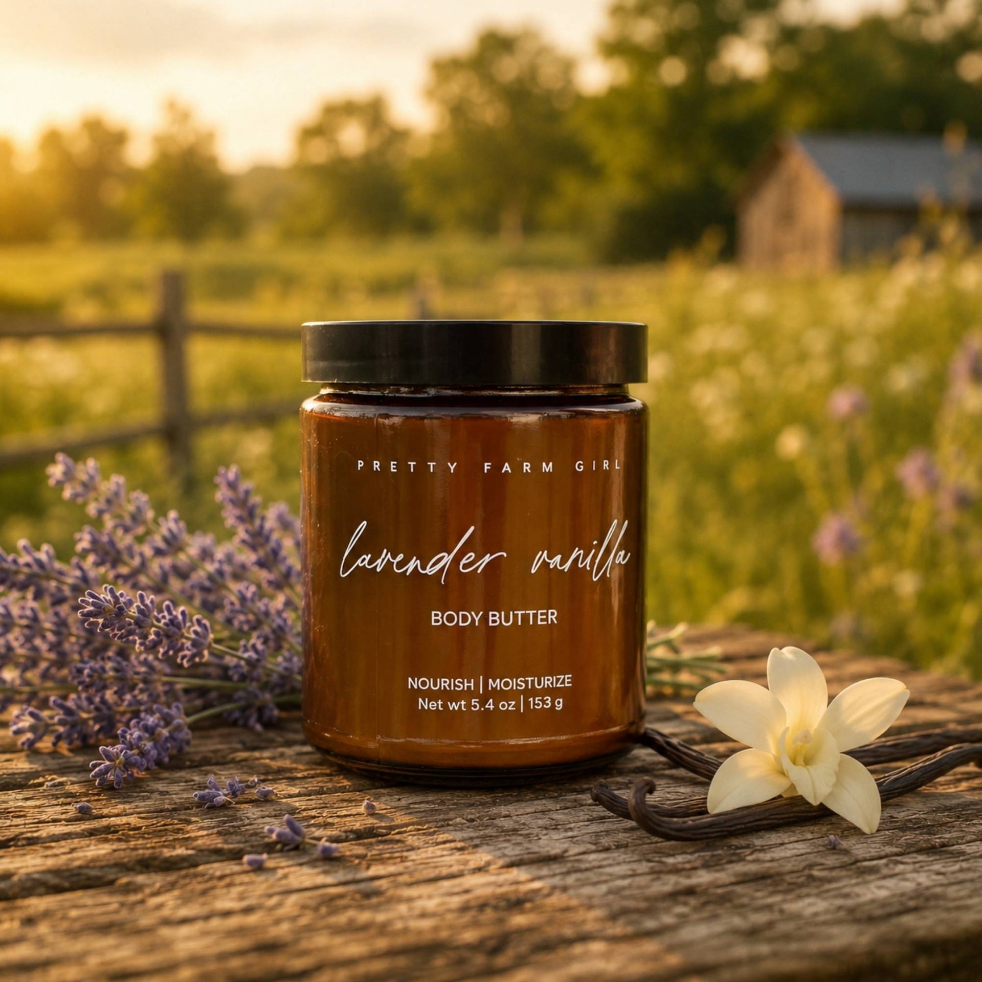 A jar of Pretty Farm Girl lavender vanilla body butter sits on a rustic wooden surface outdoors, surrounded by lavender sprigs, a vanilla bean, and a white flower, with a blurred field and fence in the background.