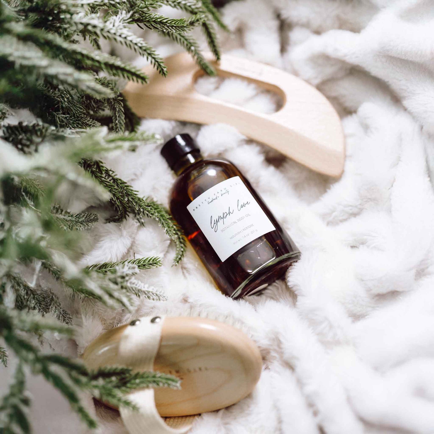 Bottle of Lymph Love body oil with wooden body gua sha and bamboo dry body brush on a white textured surface and green pine tree branches on the left side.
