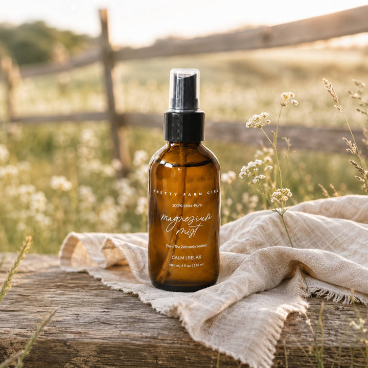 A bottle of Pretty Farm Girl body mist sits on a rustic wooden table draped with a beige linen cloth, surrounded by wildflowers and a soft-focus outdoor background.