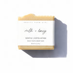 A bar of Pretty Farm Girl 'milk + honey' exfoliating face and body bar soap, wrapped in a white paper label, set against a plain white background with a soft shadow.