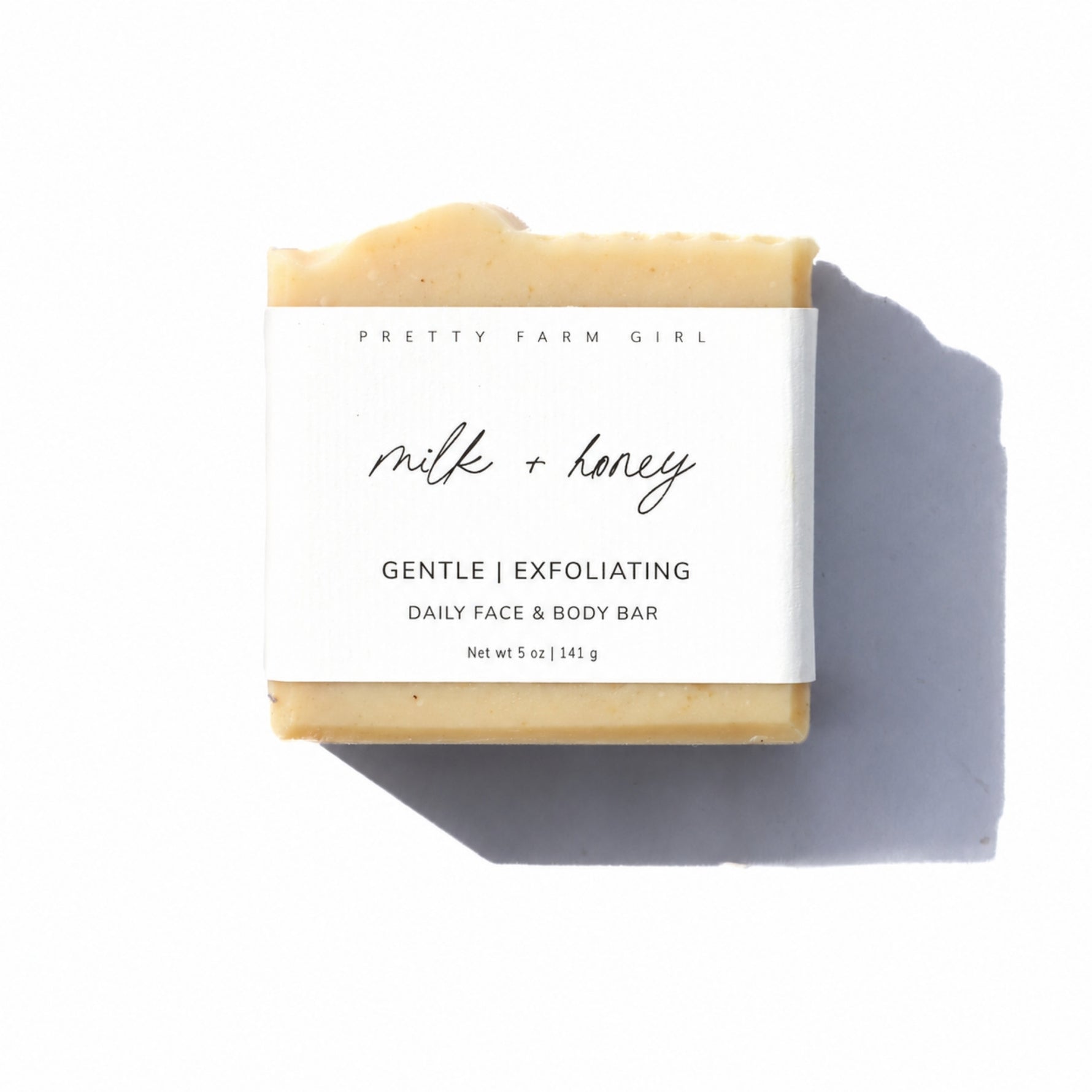 A bar of Pretty Farm Girl 'milk + honey' exfoliating face and body bar soap, wrapped in a white paper label, set against a plain white background with a soft shadow.