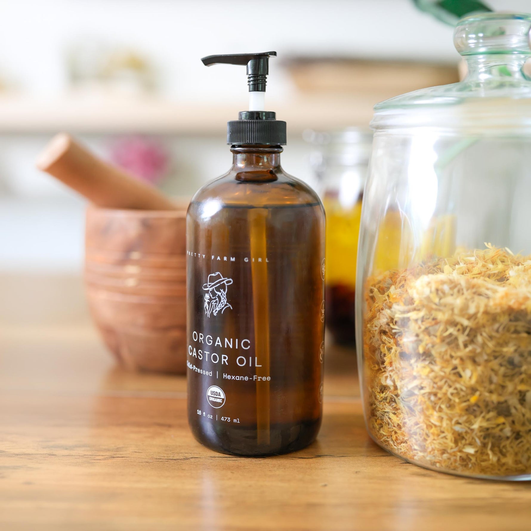 Pretty Farm GIrl's 16oz brown glass bottle of Organic Cold pressed hexane free Castor Oil on a wooden surface with a jar of herbs in the background.