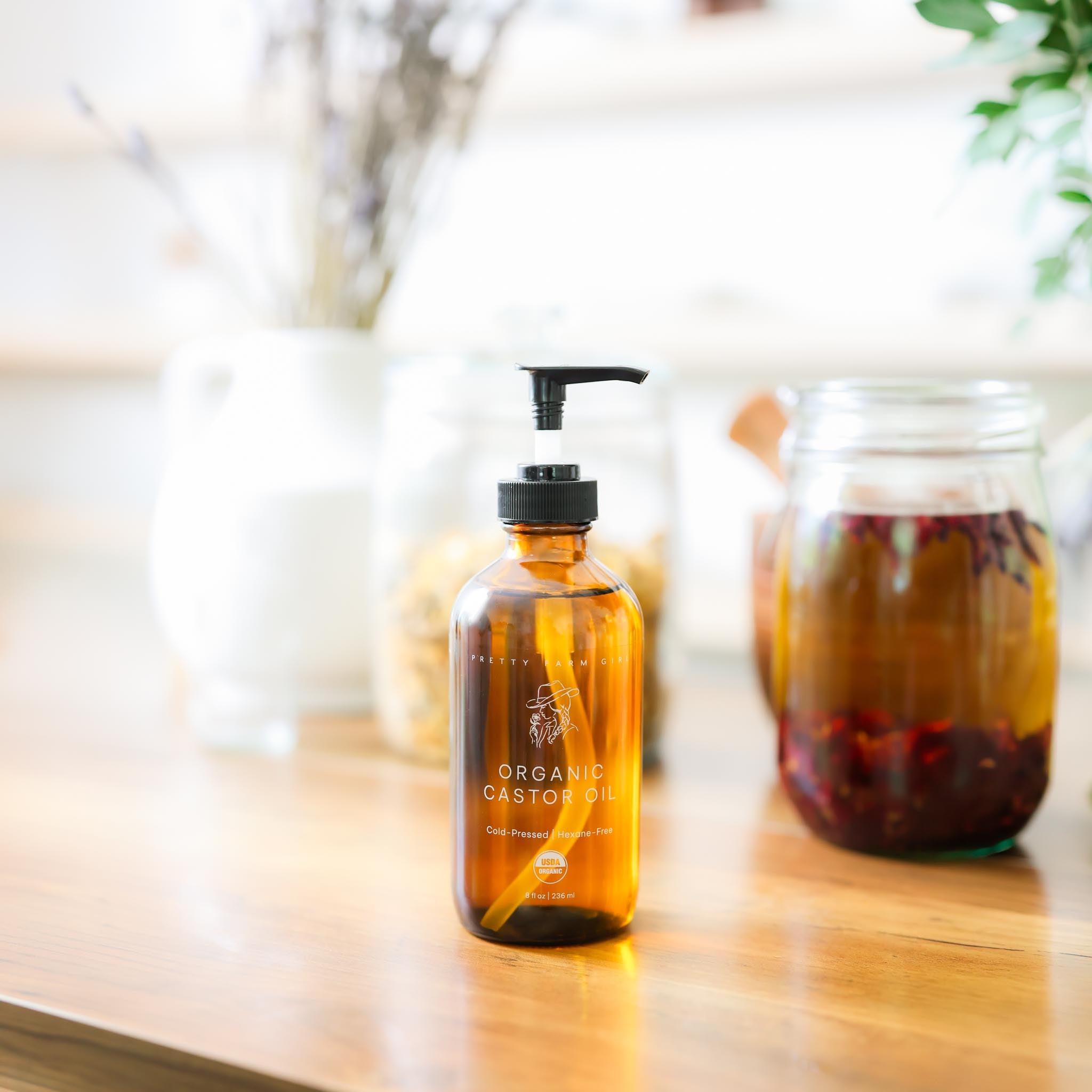 Pretty Farm GIrl's 8oz brown glass bottle of Organic Castor Oil with a pump top on a wooden surface with a jar of herbs in the background.