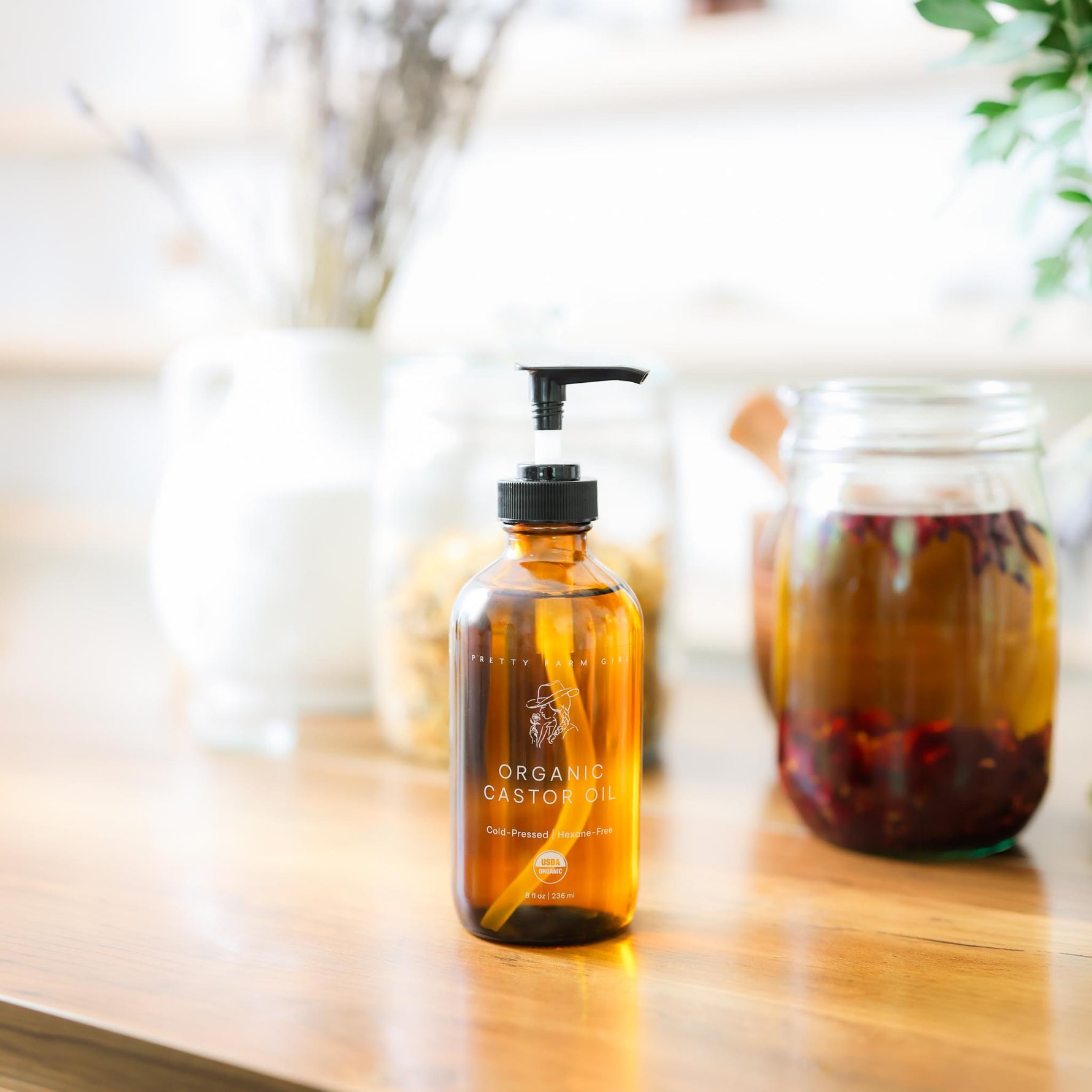 Pretty Farm GIrl's 8oz brown glass bottle of Organic Cold pressed hexane free Castor Oil with a pump top on a wooden surface with a jar of herbs in the background.