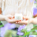 Person holding a jar of .78oz 'Original Fresh Face Cream' with blurred purple flowers in the background