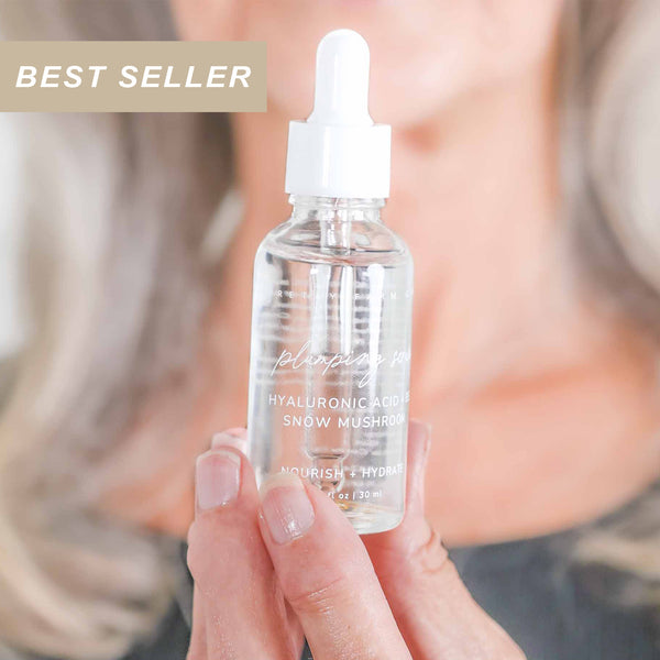 Hand holding a glass 1oz skincare dropper bottle of Pretty Farm Girl's best selling Plumping Hyaluronic acid and snow mushroom serum with a blurred background.