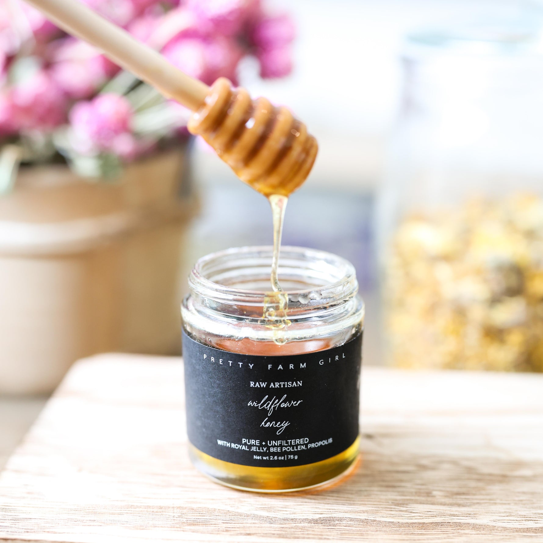 Jar of Pretty Farm Girl's Raw Artisan Wildflower Honey, Pure Unfiltered with a display wooden dipper on a wooden surface with flowers in the background.