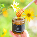 Jar of Pretty Farm Girl's Raw Artisan Wildflower Honey with a honey dipper against a natural background with flowers