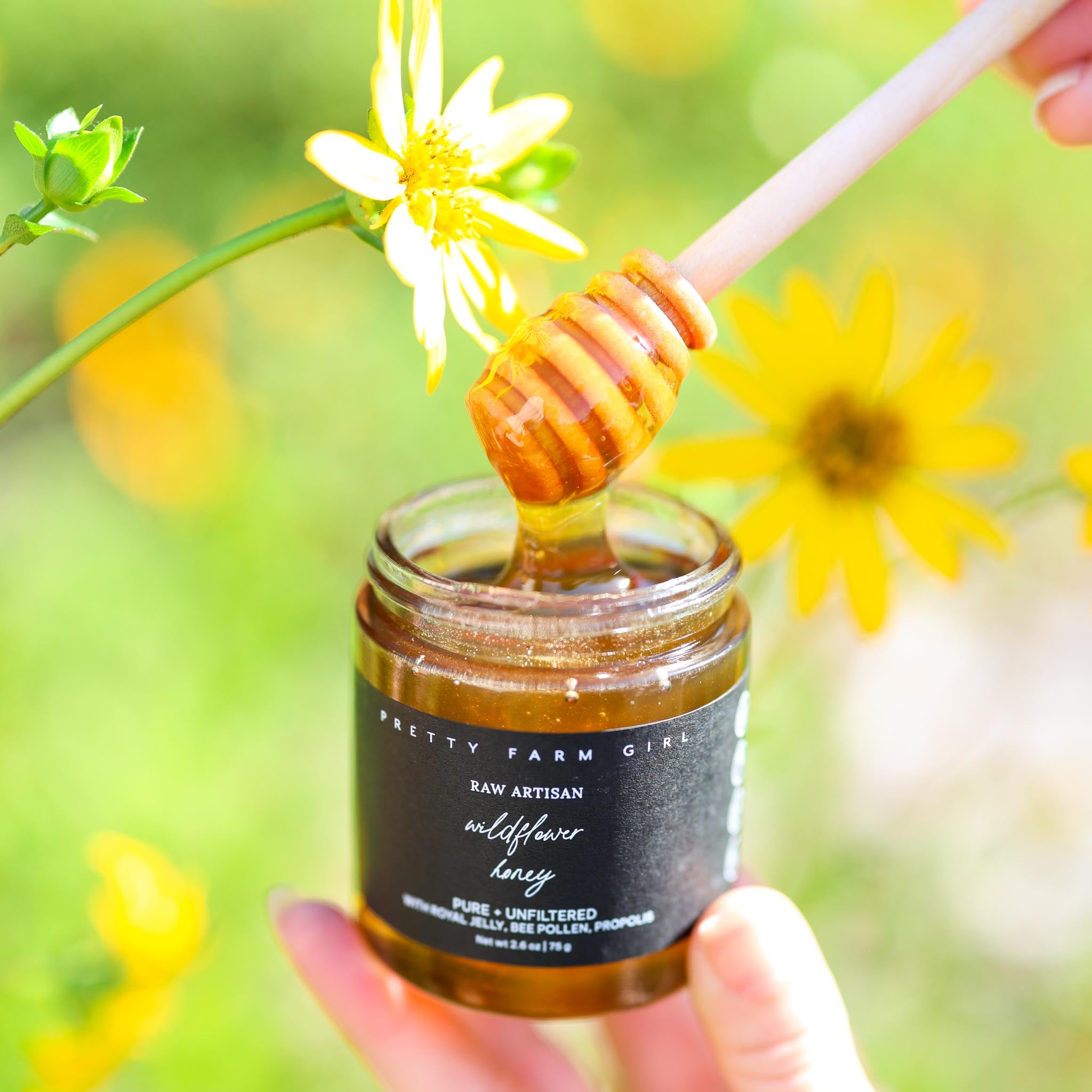 Jar of Pretty Farm Girl's Raw Artisan Wildflower Honey with a honey dipper against a natural background with flowers