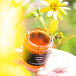 Jar of Pretty Farm Girl's Raw Artisan Wildflower Honey held in a hand with flowers in the background