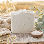 A rectangular bar of Salt + Sea soap with tiny speckles sits on a rustic wooden surface, styled with a burlap cloth, white flowers, green eucalyptus leaves, and a small wooden bowl of coarse salt.
