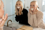 Two women applying mouth tape to their faces in front of a mirror.