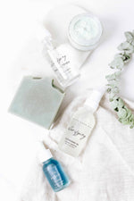 Skincare products including blue beauty tallow face cream, clarifying facial toner, blue beauty facial soap bar, clarifying cleansing oil, and blue balance serum on a white backdrop with a eucalyptus sprig.