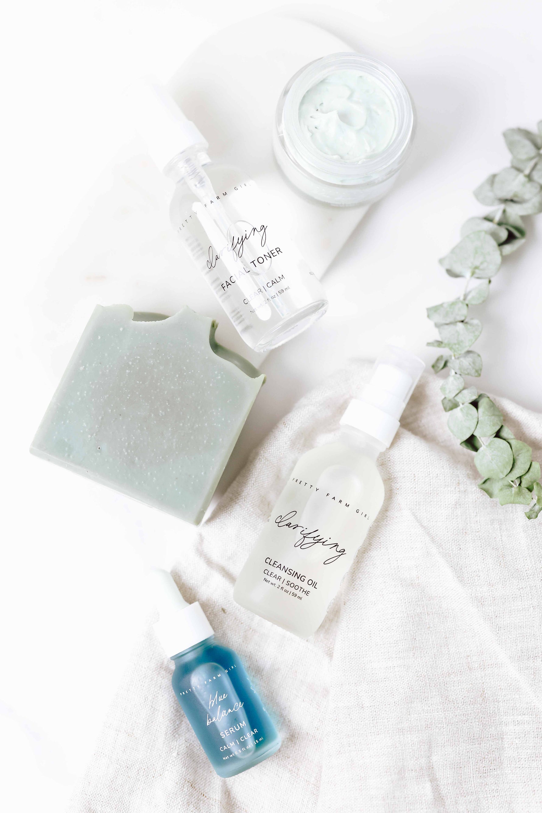 Skincare products including blue beauty tallow face cream, clarifying facial toner, blue beauty facial soap bar, clarifying cleansing oil, and blue balance serum on a white backdrop with a eucalyptus sprig.
